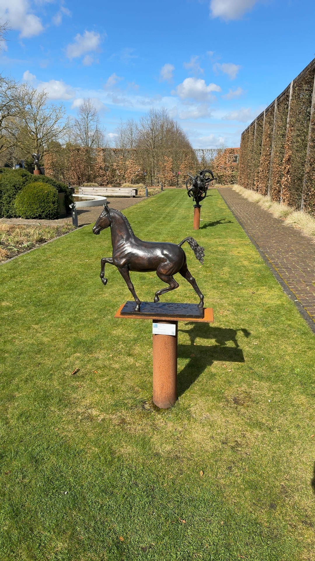 Bronze sculpture of a trotting horse by J. and M. Bremers on a rusted Corten steel pedestal in a garden. - view 1