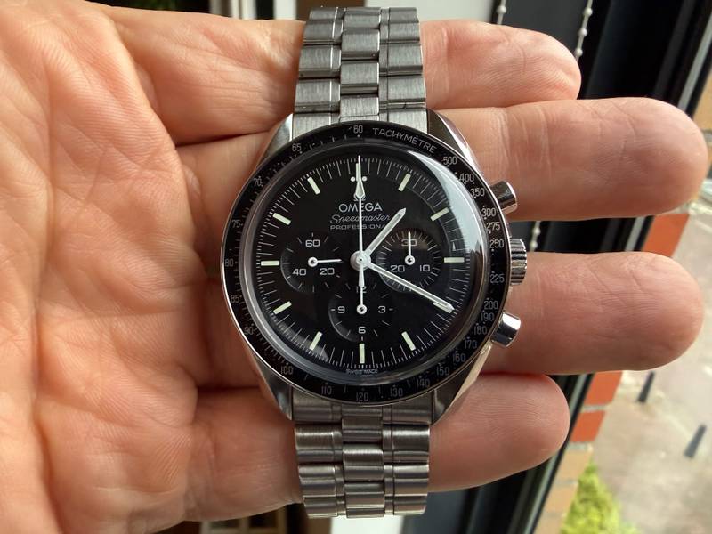 Modern Omega Speedmaster Professional with hesalite crystal, matte black step dial, and brushed five-link steel bracelet held in a hand.