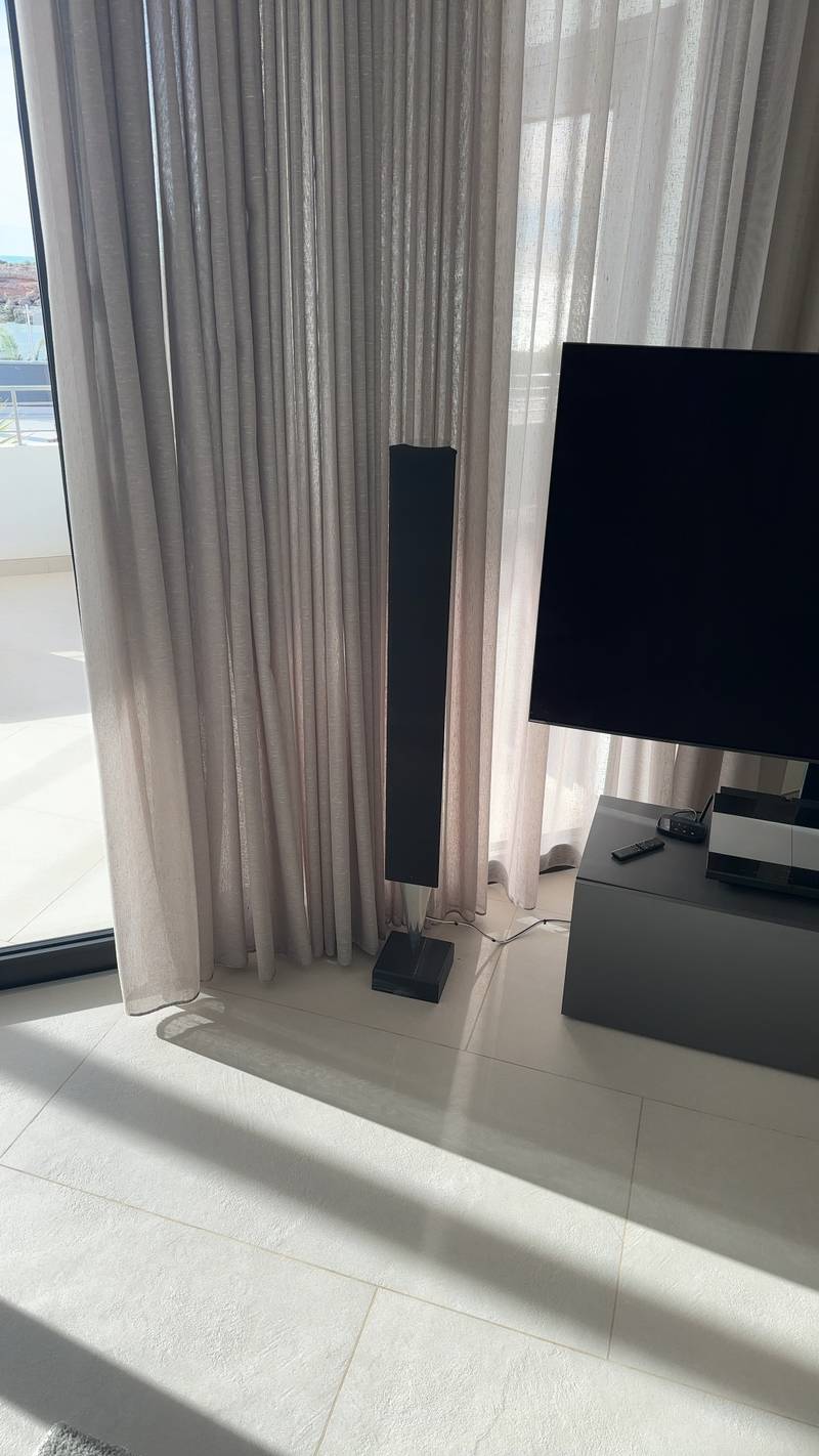 Sleek black contemporary floor-standing speaker next to a large television and light curtains in a modern room