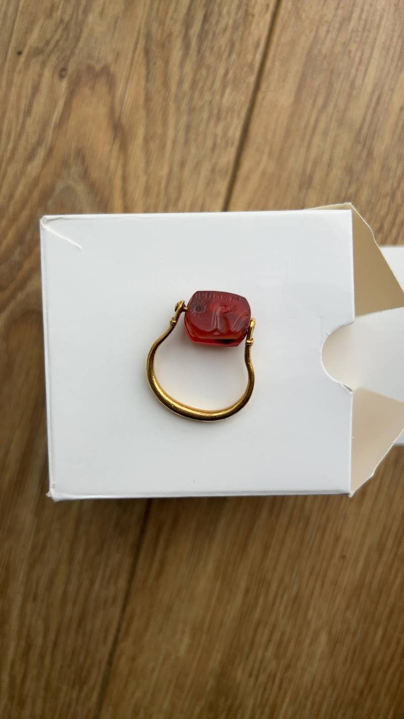 A yellow gold swivel ring with a red carnelian intaglio featuring an ancient animal carving and script, resting on a white box. - view 1