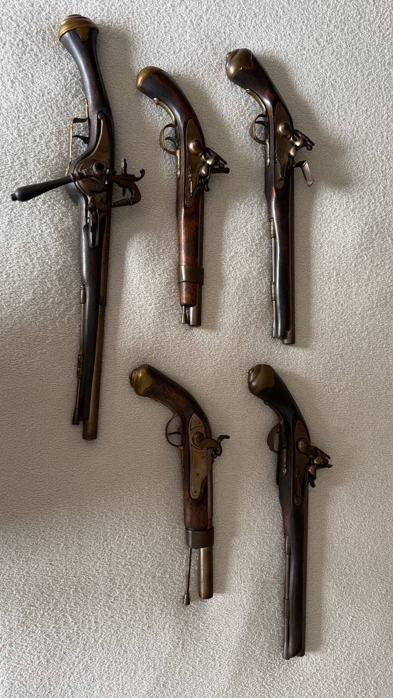 Five decorative flintlock pistol replicas with wood stocks and brass-toned metal, arranged on a textured white surface. - view 1