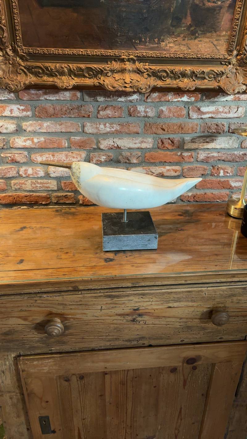White marble bird sculpture on a black marble base, placed on a wooden dresser in a room with a brick wall. - view 1