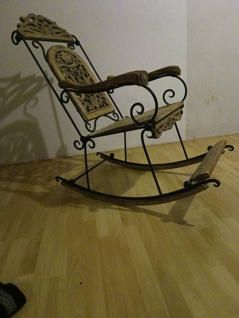 A rocking chair with a black wrought iron scrollwork frame and light-colored carved wooden panels for the seat and backrest.