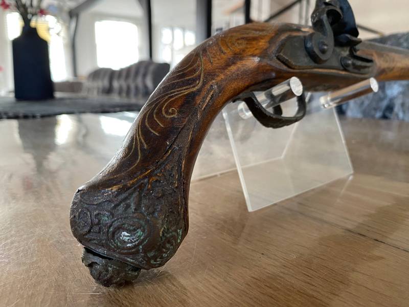Close-up of a 19th-century miquelet pistol on a display stand, showing brass wire inlay in the wooden stock and a rusted iron lock mechanism. - view 1