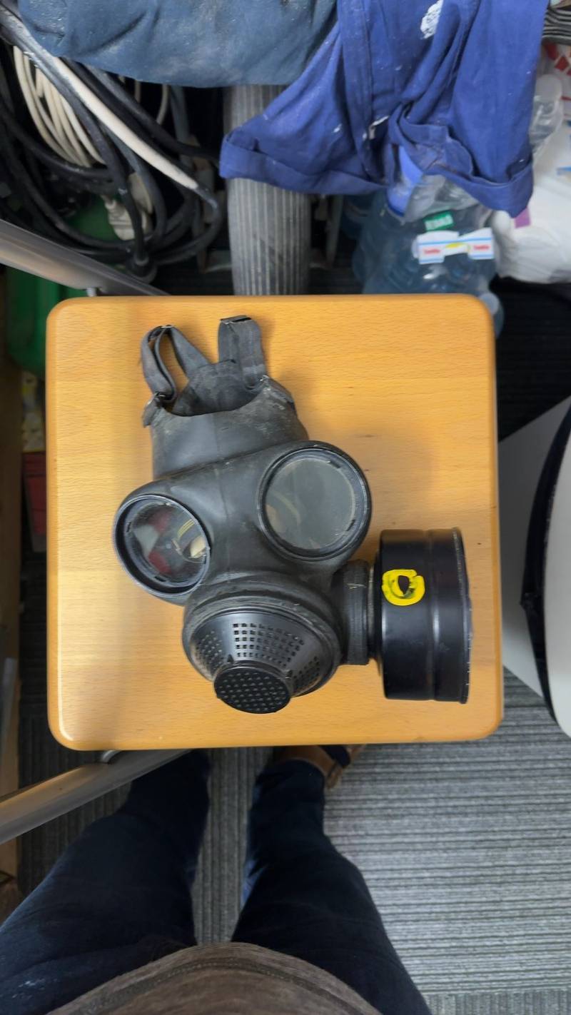 British S10 gas mask on a wooden stool with a 12/82 date stamp and handwritten harness label visible. - view 1