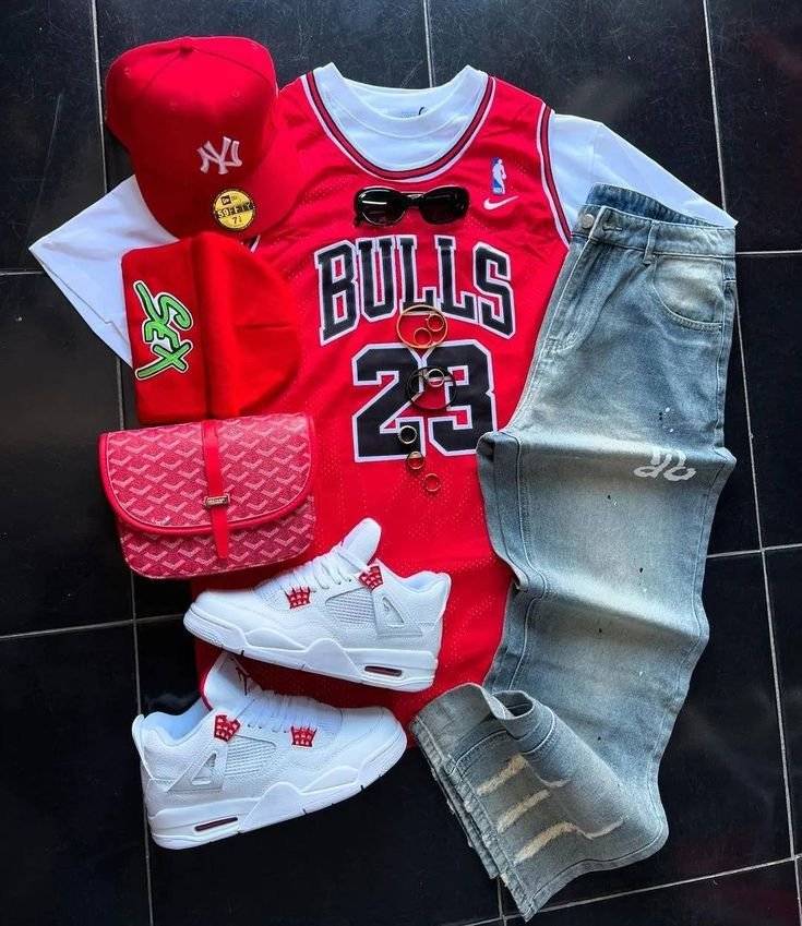 Flat lay of red Chicago Bulls jersey, white Air Jordan 4 sneakers, red patterned messenger bag, and light blue jeans on a black tile floor.