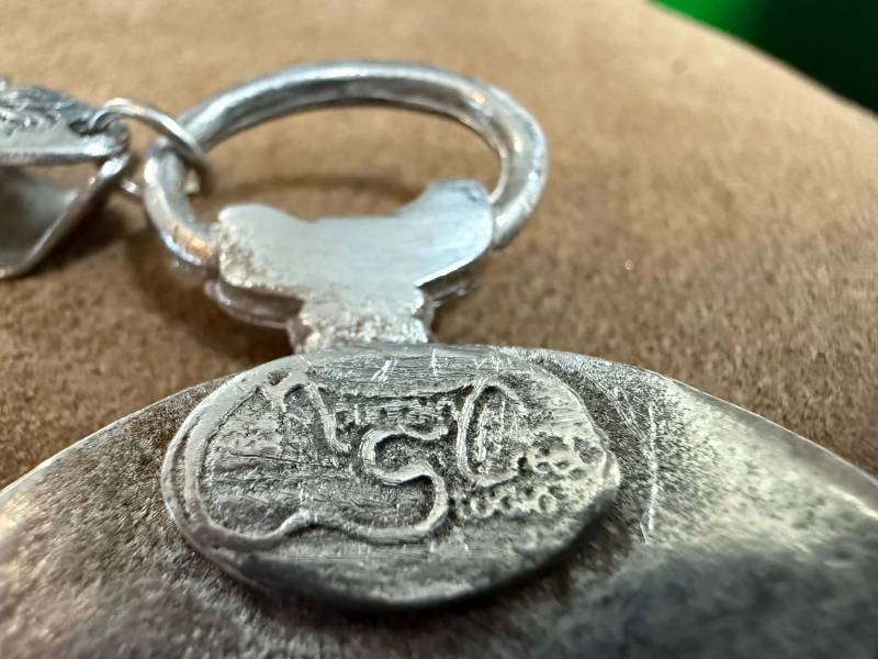 Macro photograph of a silver studio jewelry pendant reverse displaying an applied oval cartouche with raised maker initials and suspension loop.