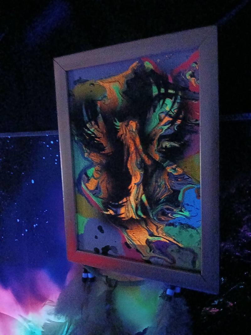 Framed abstract fluid acrylic painting glowing with orange and green pigments under ultraviolet blacklight. - view 1
